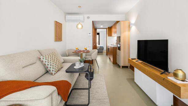 Ein modernes Wohnzimmer mit Sofa, Couchtisch und Fernseher. Im Hintergrund ist eine Küche erkennbar.