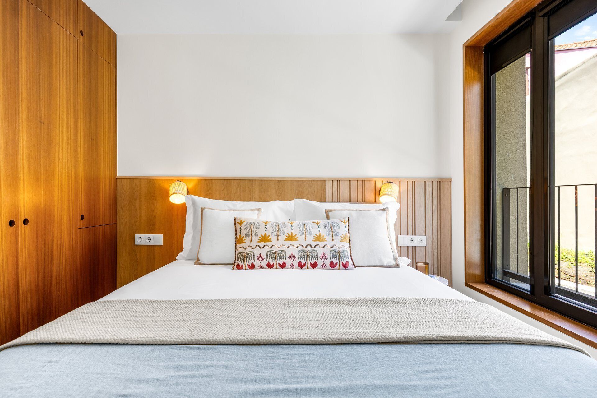 A hotel room with a large bed, wooden headboard, two bedside lamps, and a view through a large window.