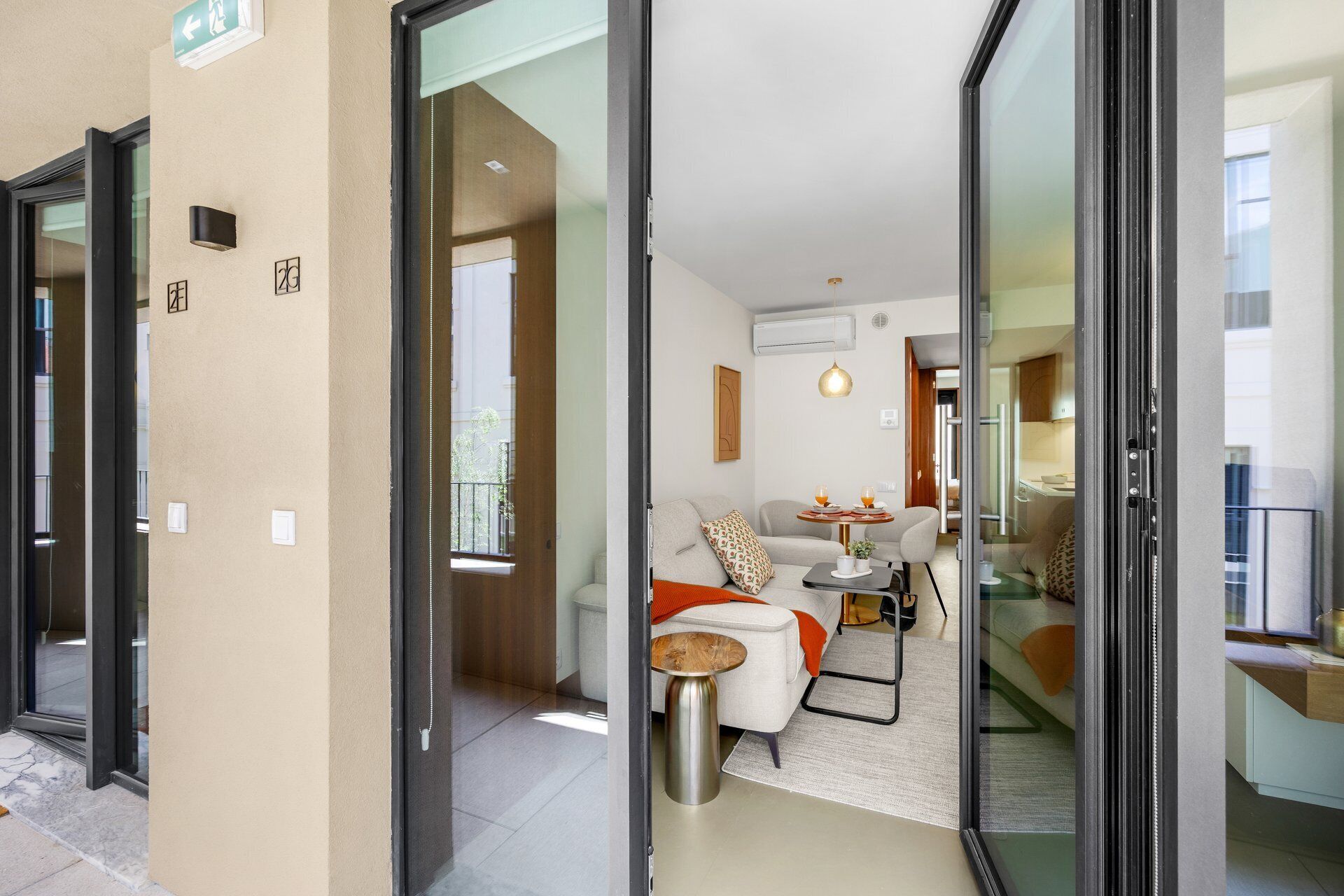 A modern apartment with a glass door entrance, a living area with a sofa, a coffee table, and a dining table with two glasses and a bottle.