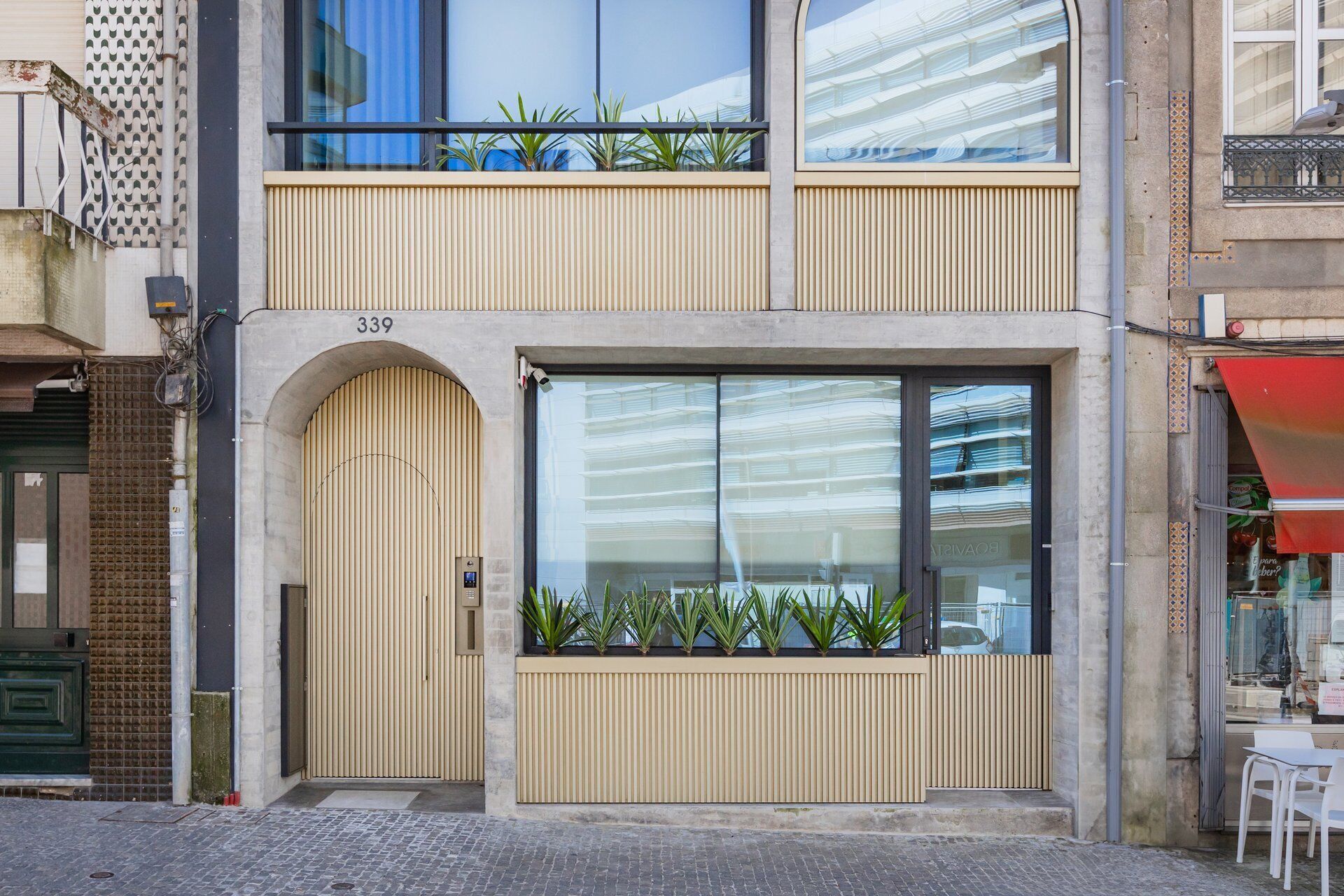 A modern building with a large window, a door, and a balcony with plants.