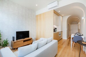A modern living room with a flat-screen TV, a light-colored sofa, wooden flooring, and a kitchen area with a dining table and chairs. - Modern 1BR with AC | Casa da Música (Porto)