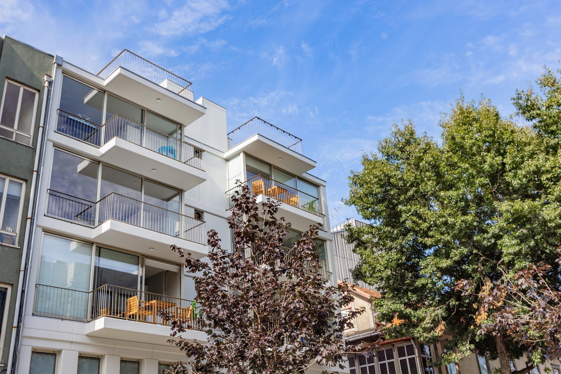 A multi-story residential building with balconies, large windows, and a tree in the foreground.