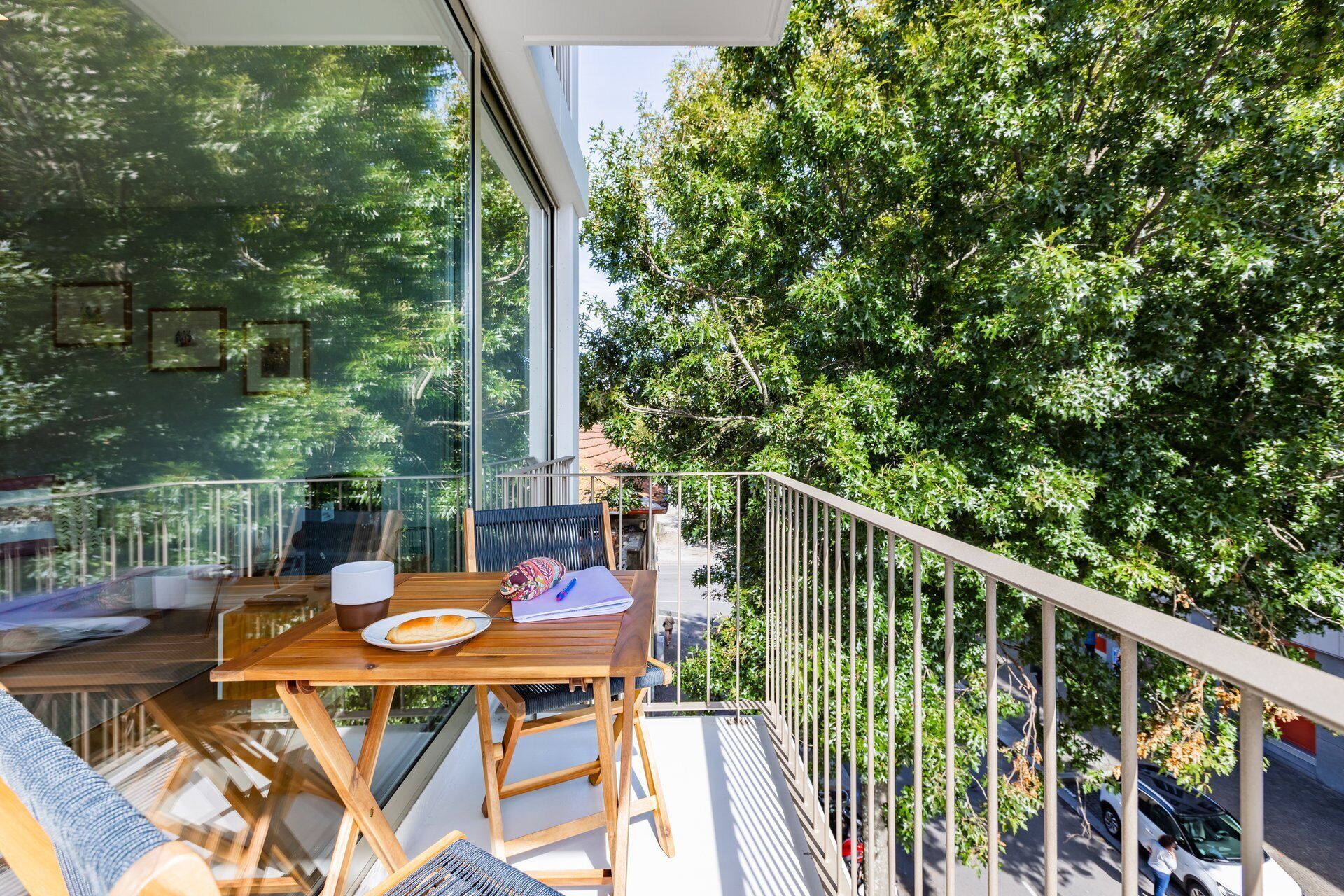 Un balcon avec une table, des chaises et une balustrade donnant sur une rue bordée d’arbres.