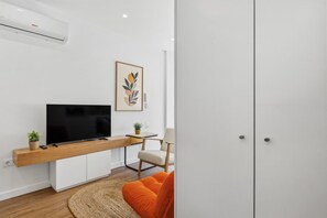 A modern living room with a flat-screen TV, a wooden TV stand, a chair, a small table with a plant, and a framed artwork on the wall. - Porto Garden | Private Patio Studio (Porto)