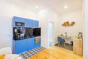 A compact kitchen with blue cabinets, a microwave, and a stove. There's a dining table with a patterned tablecloth, a blue chair, and a small desk with a lamp. - Balcony City Views | Porto (Porto)