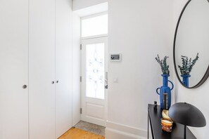 Ein Flur mit einer weißen Tür, einem kleinen Fenster, einem schwarzen Tisch mit einer Lampe und einer blauen Vase mit Pflanzen.