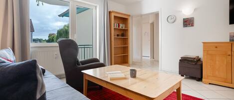 A modern living room with a sofa, a wooden table, a bookshelf, and a view through a large window.