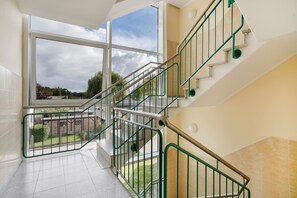 Eine Treppe mit grünen Geländern führt zu einer oberen Etage, ein großes Fenster bietet Blick ins Freie und der Boden ist mit Fliesen belegt.