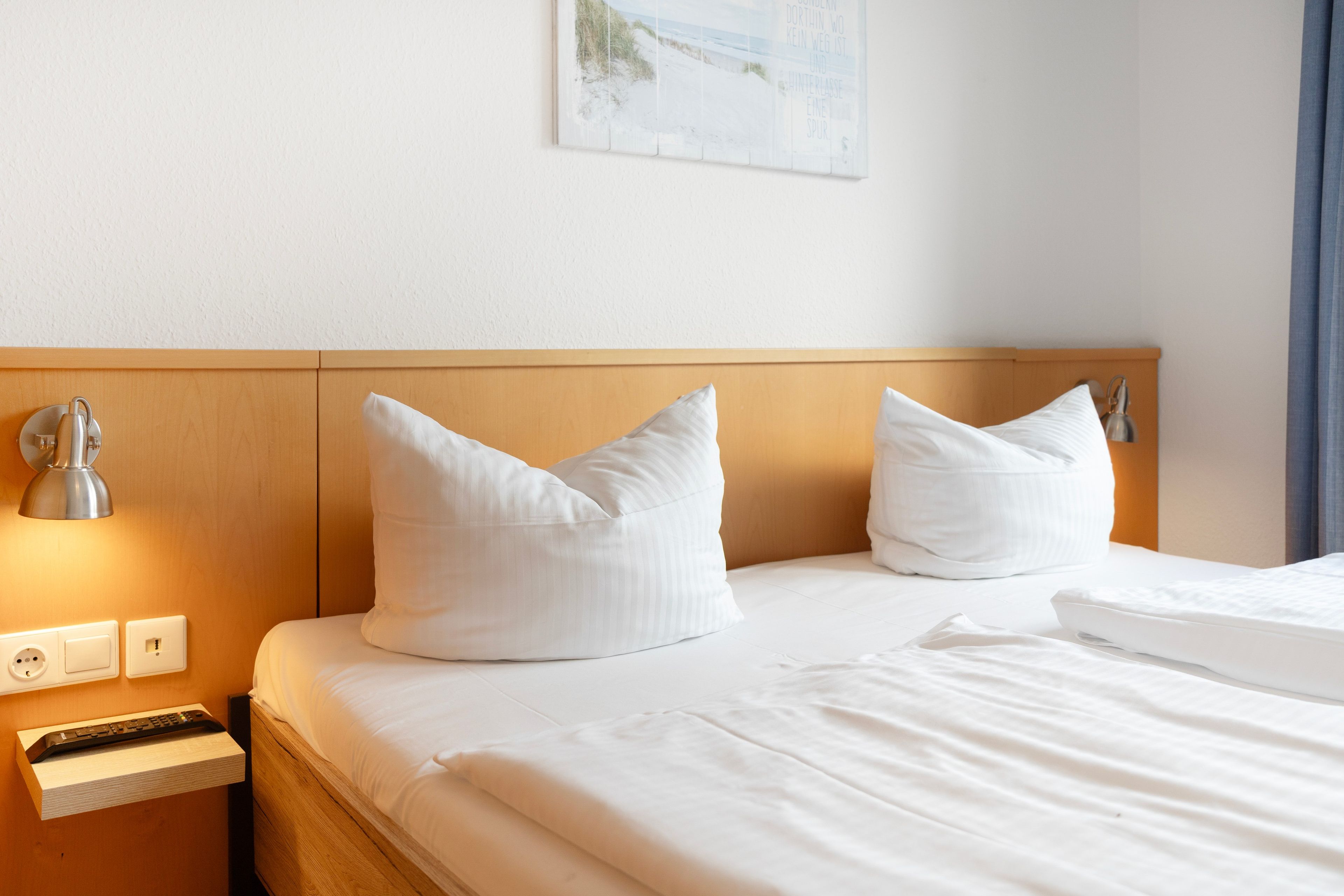 Une chambre d’hôtel avec une tête de lit en bois, deux oreillers blancs, une lampe de chevet et une table de chevet avec une lampe, ainsi qu’un tableau accroché au mur.