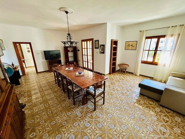 A spacious room with a large wooden dining table, a television, and a patterned floor.