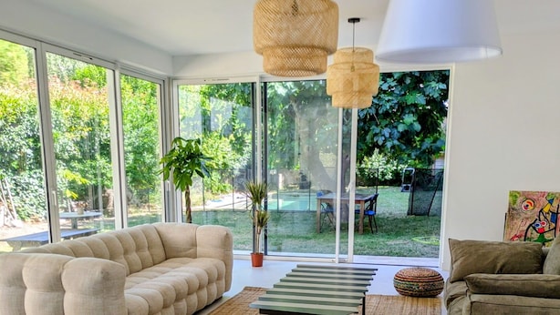 Een moderne woonkamer met een bank, poef en stoelen, grote ramen en uitzicht op de omgeving.