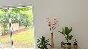 Een kamer met een grote schuifdeur naar een tuin, een wit betegelde bank met plantenpotten, en een witte muur.