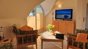 A cozy living room with a TV, floral-patterned furniture, a vase with flowers, and a view of the beach. - Apartment for 4 People (44 m²) in Zempin (Seaside Resort) (Zempin)