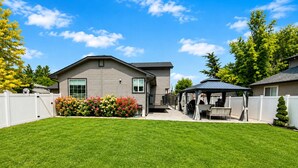 A residential backyard with a well-maintained lawn, a patio area with outdoor furniture, and a white fence. - The Hive Hideaway (Meridian)