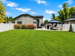 A residential backyard with a well-maintained lawn, a patio area with outdoor furniture, and a white fence. - The Hive Hideaway (Meridian)