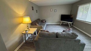 A living room with a sofa, a TV on a stand, a lamp, and a window with blinds. - Cozy Lakeside Two Bedroom Escape (Wichita)