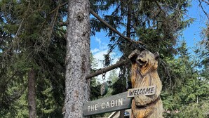Une enseigne en bois représentant une cabane, ornée d’une roue rustique et d’une figurine d’ours, le tout entouré d’arbres et se dressant sous un ciel dégagé.