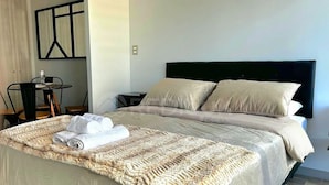 A neatly made bed with a textured blanket, a small table with a chair, and a wall art piece. - Rbk Cómodo Studio con gran ubicación (Santiago)