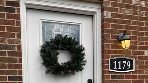 A white door with a Christmas wreath, a black house number sign, and a wall-mounted light fixture.