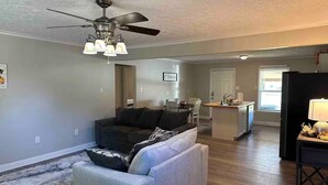 A living room with a sofa, a coffee table, a ceiling fan, and a kitchen area in the background.