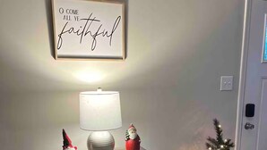 A room with a wall-mounted sign, a table with a lamp and decorative items, a small Christmas tree, and a door.