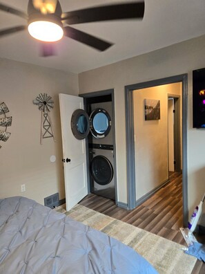 A bedroom with a bed, a ceiling fan, a washing machine, and a door leading to another room.