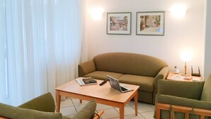 Una sala de estar con un sofá, una mesa con una computadora portátil, una silla y dos cuadros en la pared.