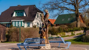 Una escultura en un parque con bancos, una casa al fondo y cielo despejado.