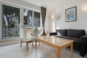 Een woonkamer met een bank, een tafel en een stoel. Er is een raam met gordijnen, een radiator en een schilderij aan de muur.