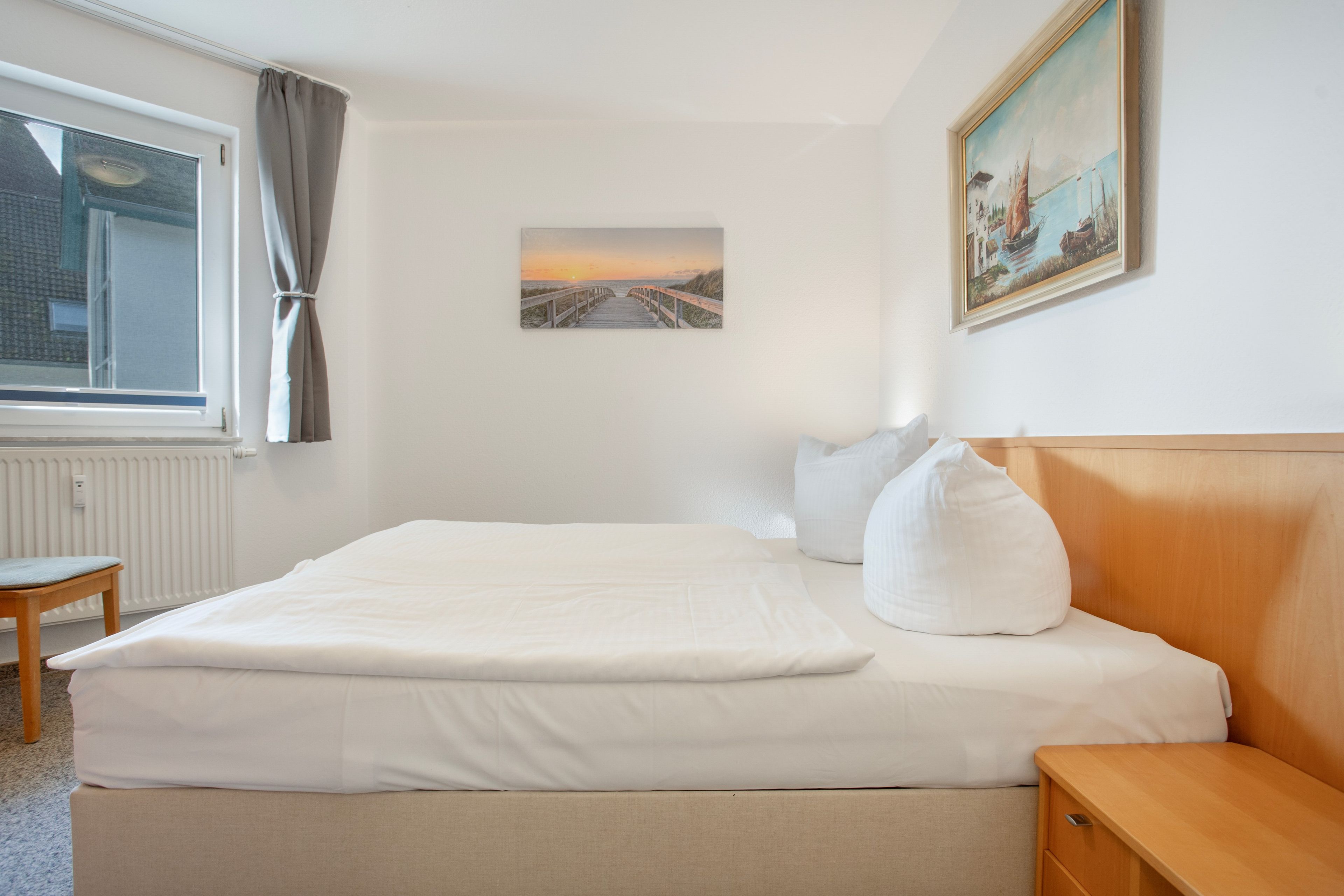 A hotel room with a bed, a wooden nightstand, a chair, a window with curtains, and two paintings on the wall.