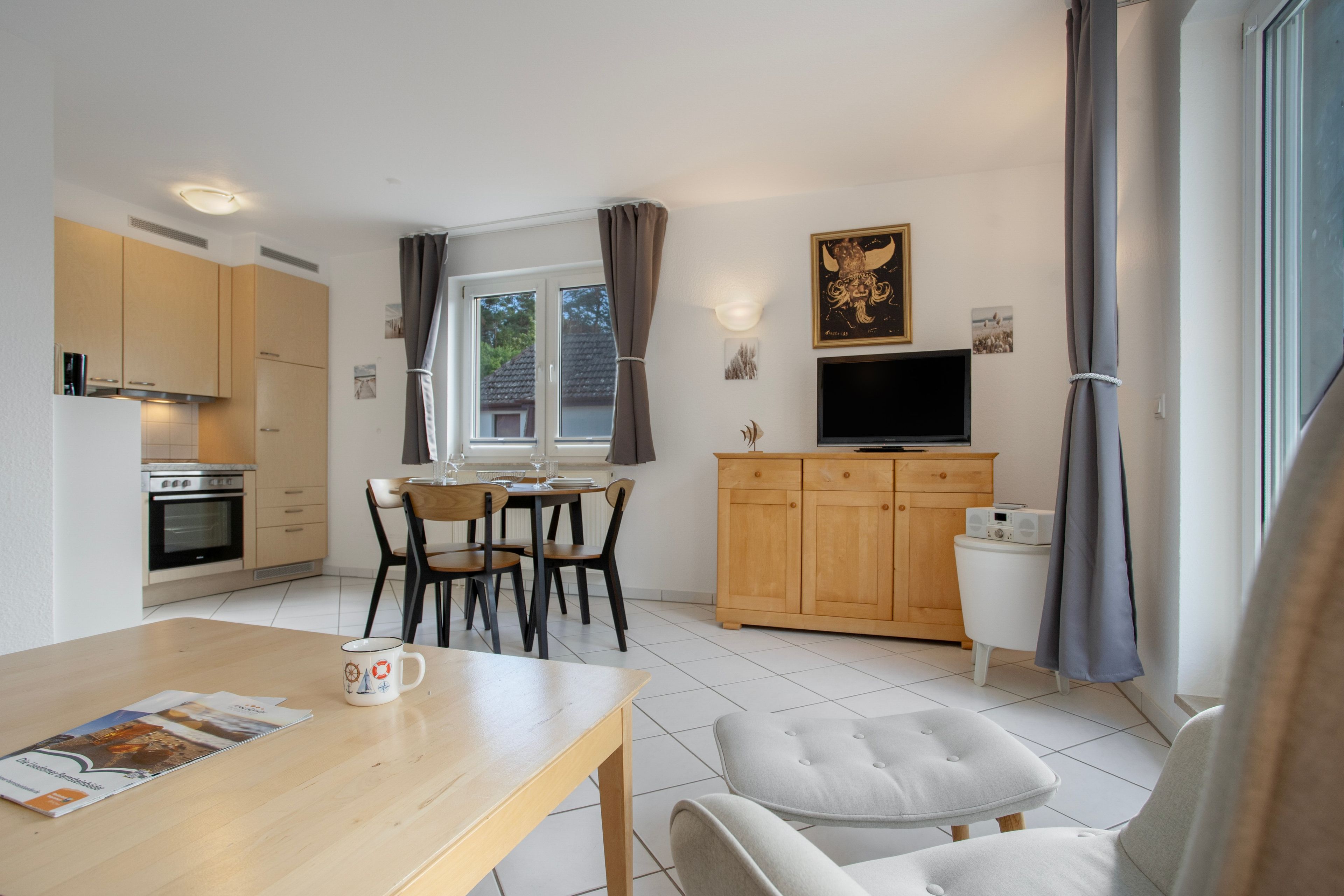 A compact kitchen with modern appliances, a dining area with a table and chairs, a television mounted above a wooden cabinet, and a window with curtains.