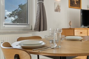 A dining table set with plates, glasses, and cutlery, a window with a view of trees, and a television on a cabinet. - Apartment 'Möwe' with Balcony and Wi-Fi (Zempin)