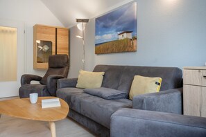A modern living room with a grey sofa, a wooden coffee table, a chair, and a painting of a beach house. - Apartment "Bernsteinfischer" with Balcony and Wi-Fi (Zempin)