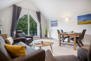 Ett modernt vardagsrum med ett stort fönster, en matplats med ett träbord och stolar samt en mysig sittgrupp med en soffa och fåtöljer.