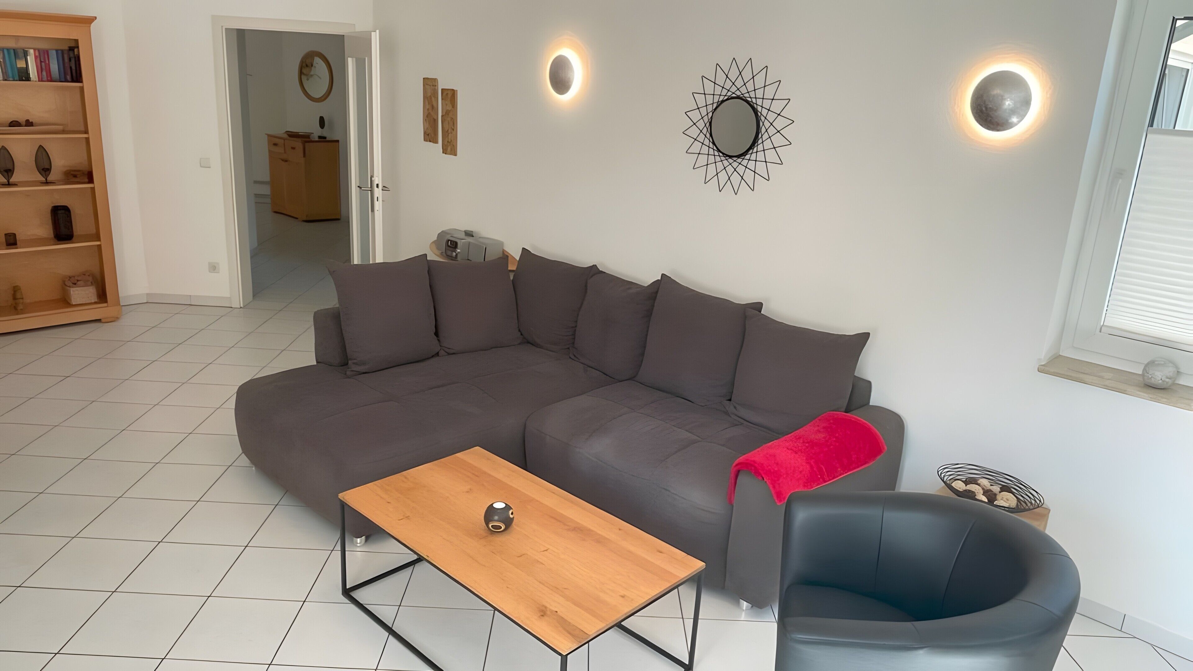 Een woonkamer met een hoekbank, een salontafel, een zwarte fauteuil en een boekenkast.