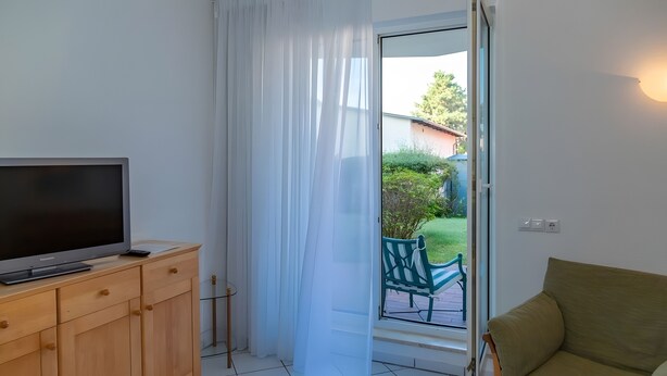 Una stanza con televisore su un mobile in legno, una porta scorrevole in vetro che dà su un balcone e una sedia verde all'esterno.