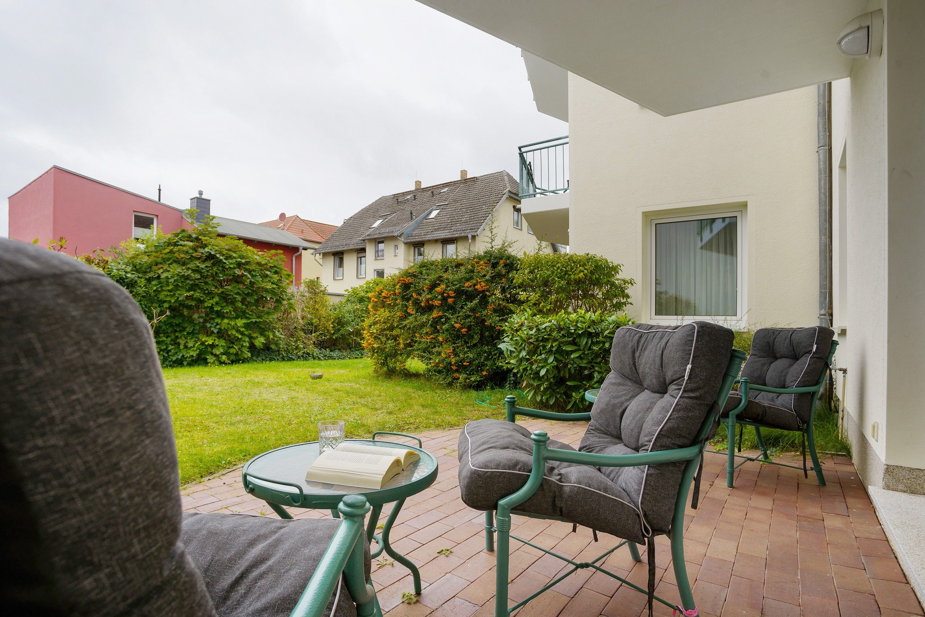 An outdoor patio with two chairs and a table, a garden, and a building in the background.