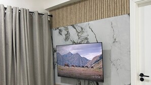 A modern living room with a flat-screen TV mounted on a wall with a marble-like texture, a wooden panel design, and a dark wood TV stand with drawers. - Cozy 1BR Apt in Hulhumale, Maldives - AC/WiFi (Hulhumale)