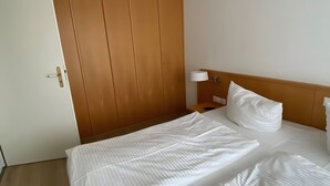 Hotellihuone, jossa on puinen vaatekaappi, yöpöytä lampun kanssa ja siististi pedattu sänky valkoisilla vuodevaatteilla.
