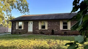 A single-story brick house with a front door, two windows with shutters, and a small front yard.