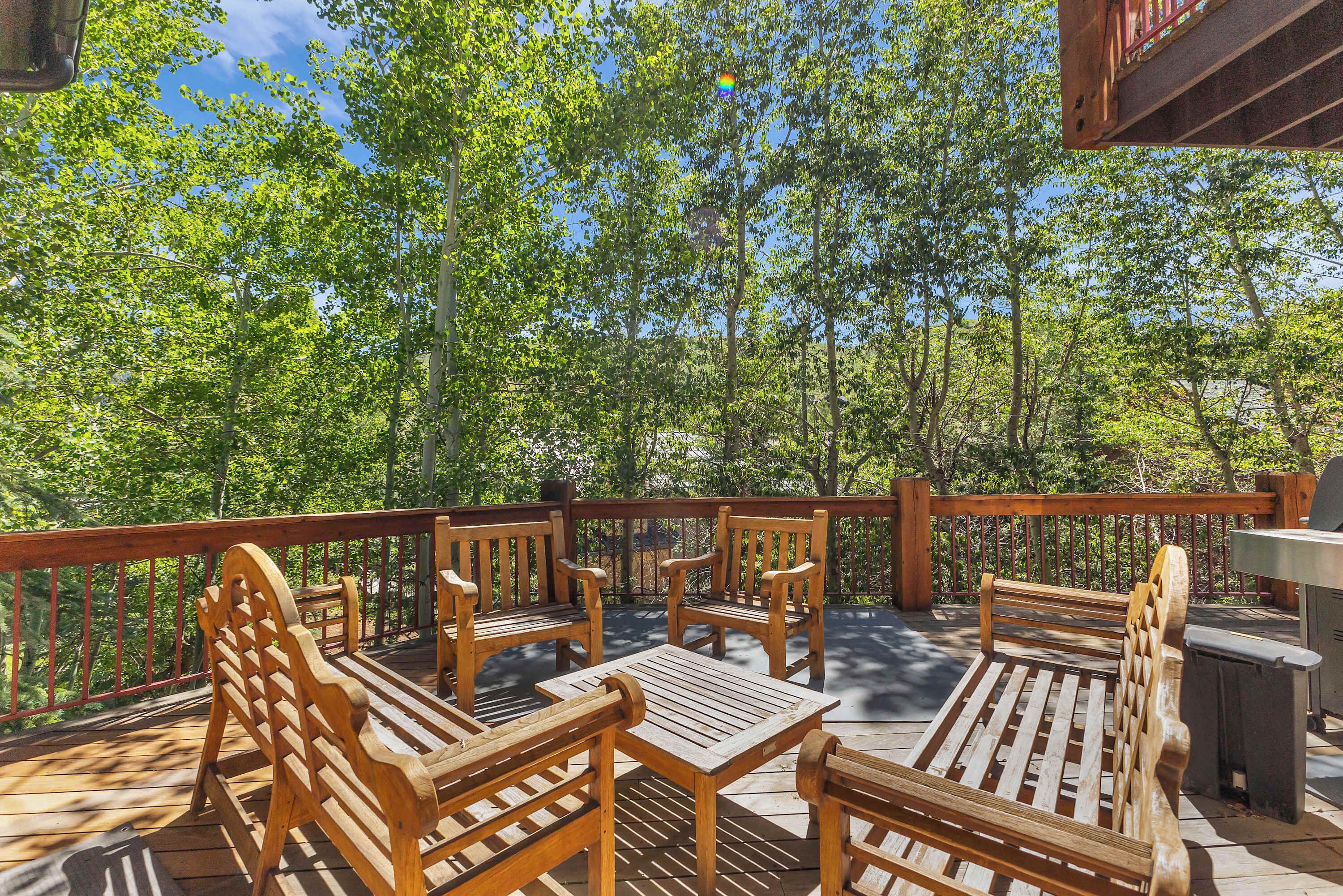 A wooden deck with benches and a grill area, surrounded by trees.