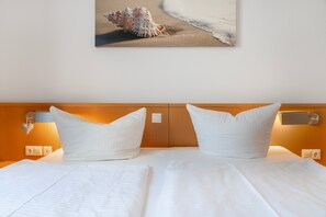 Ein Hotelzimmer mit einem Bett, zwei Kissen, einem Nachttisch, einer Wandlampe und einem Bilderrahmen mit einer Meeresschnecke an der Wand.