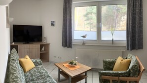 A living room with a flat-screen TV, wooden furniture, a window with a view of trees, and a radiator. - Apartment Ariane with Wi-Fi (Zempin)