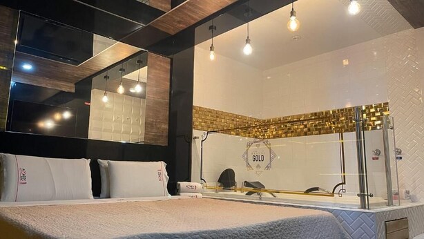 Habitación de hotel con cama, un espejo grande, ducha con pared de azulejos dorados e iluminación colgante.