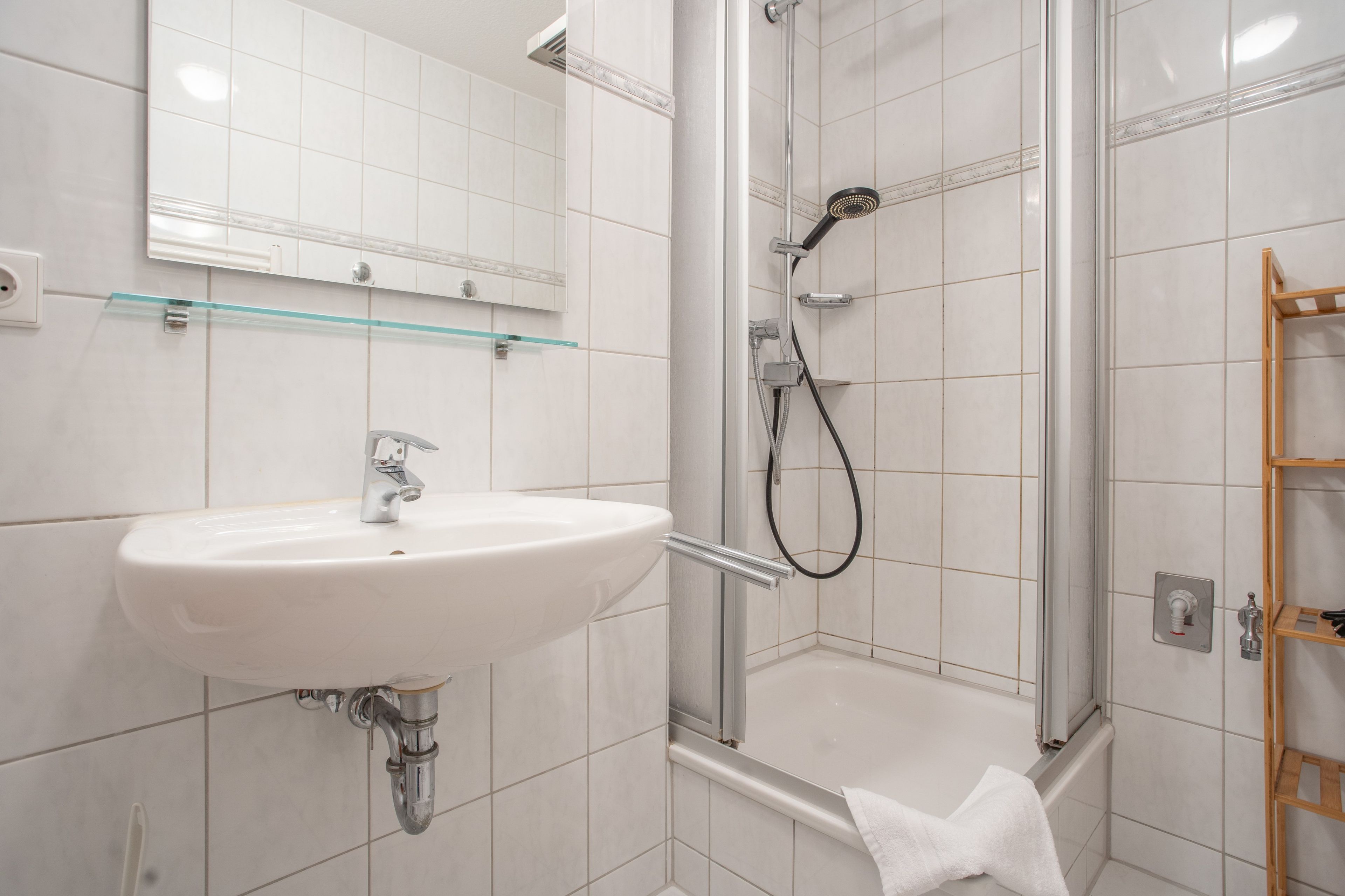 A bathroom with a white sink, a glass shower enclosure, and a wooden shelf.