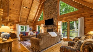 A log cabin interior with a stone fireplace, wooden furniture, and large windows. - Ripple Creek Lodge | Mtn Cabin & Firepit (Cullowhee)