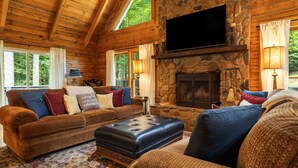 A log cabin interior with a stone fireplace, a flat-screen TV mounted above it, and a cozy seating area with a patterned rug. - Ripple Creek Lodge | Mtn Cabin & Firepit (Cullowhee)