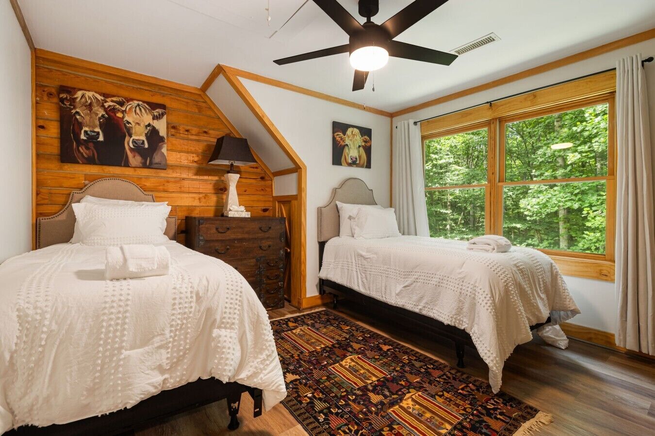 A bedroom with two beds, a ceiling fan, a large window, and a wooden wall with artwork.