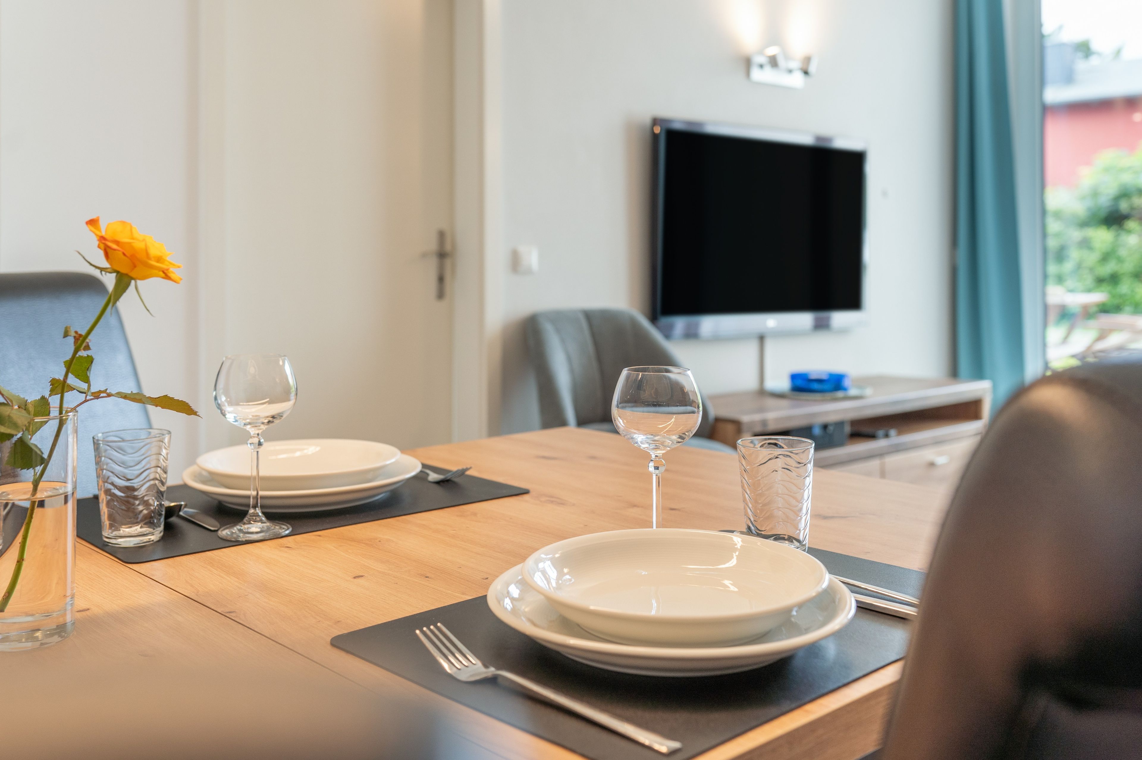A dining table set with plates, glasses, and cutlery, a rose in a vase, and a television in the background.