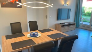 A modern dining area with a wooden table, black chairs, a blue centerpiece, a TV on the wall, and a view of greenery outside. - Apartment “Strandnah” with Private Terrace and Wi-Fi (Zempin)